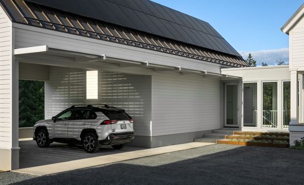 Exterior view of a contemporary residential building with a covered carport, dark standing-seam pitched roof, light horizontal cladding, and a glazed entry connection. A vehicle is parked beneath the carport, with gravel paving and small entry steps in the foreground.