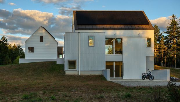 Exterior view of a contemporary two-storey house with pale vertical cladding, a steep asymmetrical roof incorporating a large dark solar-panel array, and a recessed glazed balcony or entrance zone. A separate simple gable-roofed wing stands behind it on a gently sloping site surrounded by trees.