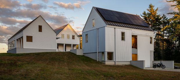 Exterior view of a small cluster of contemporary white-clad gabled houses on a gently sloping site. The buildings feature vertical and horizontal light-coloured cladding, dark pitched roofs with prominent solar panels, simple rectangular windows, and partially elevated lower levels. Trees, lawn, driveway, and a bicycle surround the residences under a partly cloudy sky.