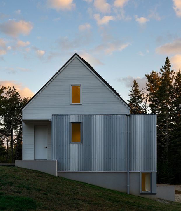 Exterior view of a compact, steep-gabled house set among trees on a sloping site. The facade combines pale horizontal weatherboard cladding with a contrasting corrugated metal-clad projecting volume, dark roof edges, simple rectangular windows with warm amber glazing, and a recessed entry porch. The image has a quiet contemporary residential character under a partly cloudy sky.