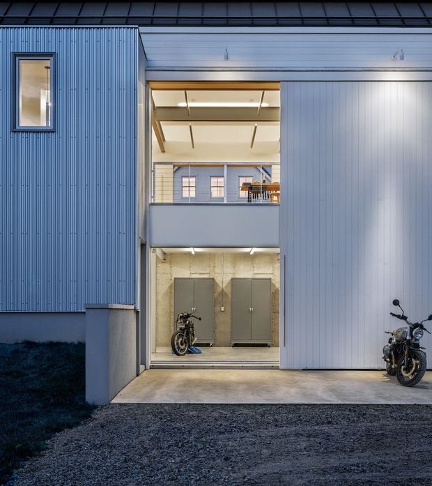 Exterior view of a contemporary multi-level residential building with blue-grey vertically ribbed cladding and a large open garage or entry bay. Two motorcycles are parked in the covered ground-level area and on the driveway. The opening reveals a concrete-finished lower level, an illuminated upper-level balcony or circulation space, and exposed ceiling beams. The scene is photographed at dusk with cool exterior lighting and warm interior illumination.