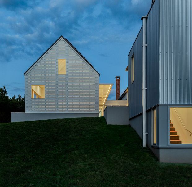 Twilight exterior view of a contemporary residential building on a sloped grassy site. The composition shows two adjacent or connected buildings with steep gabled and angular roof forms, dark grey-blue corrugated metal cladding, visible downpipes, and warm light glowing through strategically placed windows. A glazed lower-level opening reveals an interior stair, while a central covered connection and brick chimney add architectural detail.