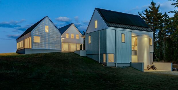 Exterior view of a contemporary multi-building residential compound at dusk. Three steeply gabled, metal-clad buildings sit on a sloping grassy site, with corrugated or ribbed light-coloured facade panels, dark pitched roofs, and warm light glowing from windows and glazed openings. The foreground building has a tall, partially glazed ground-level opening and a small bicycle near the entrance; mature trees frame the right side.