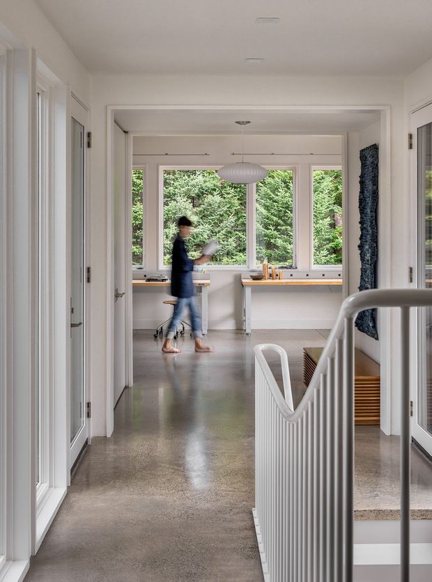 A bright, minimalist residential hallway or landing opening into a compact study or work area. The space features polished grey concrete flooring, white walls and doors, extensive garden-facing windows, a long built-in desk with timber edging, a sculptural pendant light, and a white vertical-bar stair balustrade in the foreground. A person is walking through the room, while greenery is visible outside the glazing.