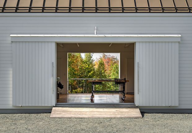 Exterior view of a contemporary light-grey vertically clad outbuilding or garage with large sliding doors opened to reveal an interior workspace or storage area. The opening frames a leafy landscaped setting, while a dark metal overhead roof structure and gravel foreground reinforce the utilitarian architectural character.