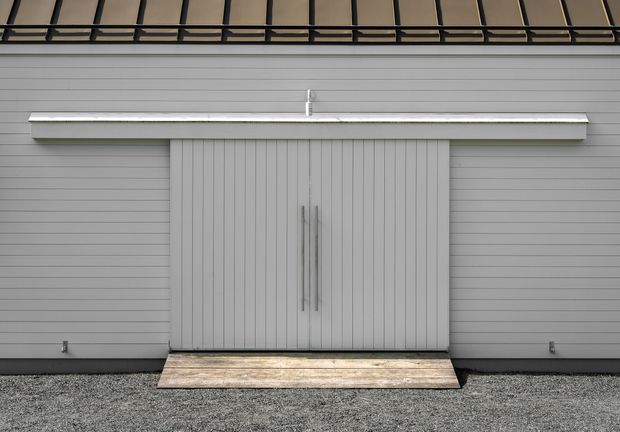 Front-facing exterior detail of a contemporary garage or storage building, featuring light grey horizontal cladding, a central vertically grooved double door with slim metal handles, a projecting canopy, and a dark exposed roof structure above. A small timber threshold sits against a gravel forecourt.