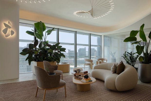 A refined high-rise living room with expansive floor-to-ceiling glazing and city views. The space features a sculptural curved cream sofa, upholstered lounge chairs, rounded wood coffee tables, a large striped area rug, abundant indoor plants, and a striking contemporary radial pendant light. Neutral warm tones and curved forms create an elegant, softly luxurious atmosphere.
