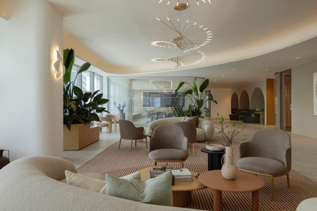A spacious, refined contemporary living room with a curved architectural layout, neutral upholstered seating, layered round coffee tables, abundant indoor plants, and a large striped area rug. Floor-to-ceiling glazing opens the room toward a bright enclosed balcony or sunroom, while sculptural ring pendant lights provide a distinctive focal point. Built-in arched niches and warm timber joinery are visible toward the rear.