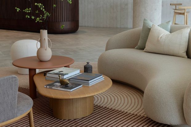 A warm, refined contemporary living room featuring a large curved beige sofa, sculptural light lounge chair, layered round timber coffee tables, neutral cushions, books, and ceramic decor. A striped curved-pattern rug anchors the seating area, with a dark timber built-in wall and textured pale wall finishes in the background.