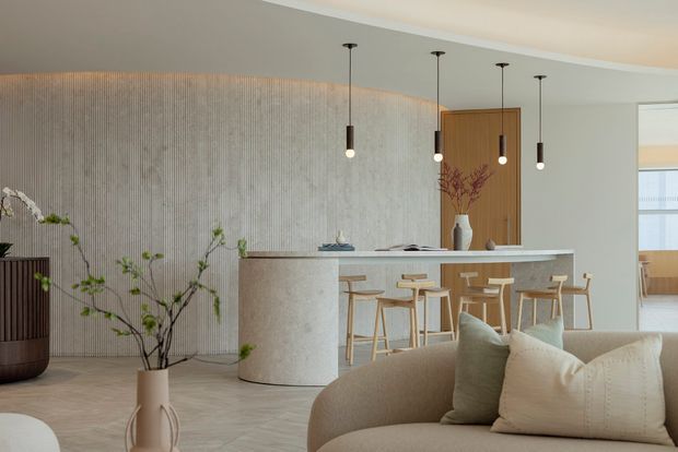 A refined open-plan kitchen and living area with a curved island featuring a pale stone or terrazzo finish, light timber cabinetry, and a textured vertical wall surface. Four slim pendant lights hang above the island, while timber bar stools and a neutral curved sofa create a warm, minimalist entertaining space. A timber door and glimpse of an adjoining room are visible in the background, with restrained natural branch decor.