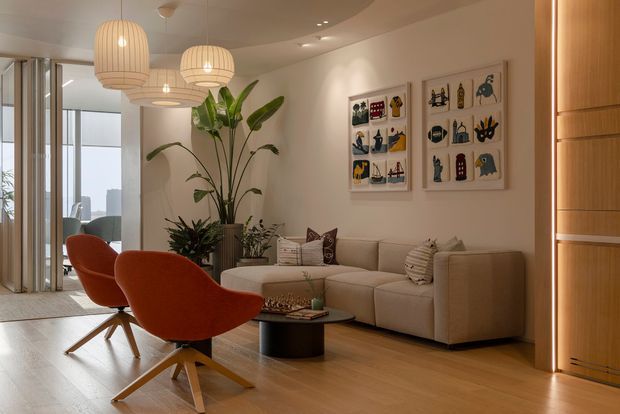 Warm, refined contemporary living room with a large beige sectional sofa, two sculptural rust-orange lounge chairs, a low dark coffee table, timber flooring, and decorative pendant lighting. A curved ceiling detail and recessed lighting add architectural interest, while framed colourful artwork and indoor plants provide accents. Full-height glazing and a timber-lined doorway are visible at the edges of the room.