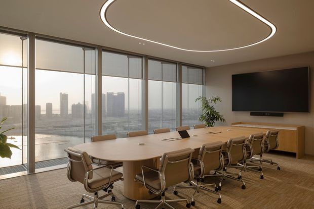 Contemporary high-rise meeting or boardroom with a long rounded rectangular conference table, numerous tan upholstered rolling chairs, a wall-mounted display, low timber media cabinetry, and floor-to-ceiling glazed windows overlooking a dense city skyline and water. A sculptural linear LED ceiling light and indoor planting add a refined commercial interior character.