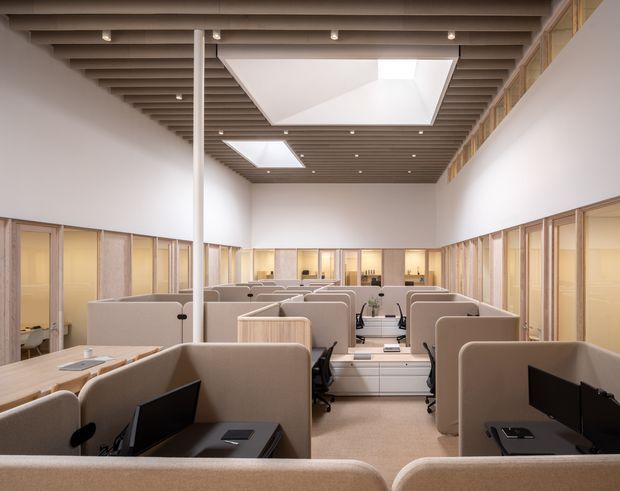 An open-plan commercial office interior viewed from an elevated position, with rows of upholstered workstation partitions, desks, monitors, and integrated storage. Glazed meeting rooms or private offices line both sides, while a warm timber slatted ceiling with recessed spotlights and large rectangular skylights creates a bright, refined workspace. The palette is predominantly white, beige, taupe, and light timber.