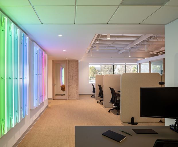 Contemporary open-plan office interior with rows of workstations separated by tall rounded acoustic screens. The space features exposed ceiling services, suspended grid ceiling panels, extensive glazing, warm carpet flooring, and a striking linear wall installation with multicoloured illuminated panels.