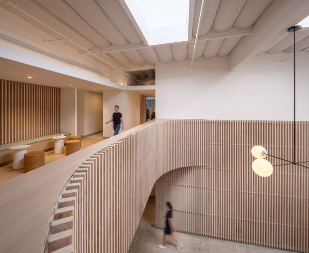 A contemporary multi-level interior with a sweeping curved mezzanine or stair balustrade clad in vertical light timber battens. The open void is lined with matching slatted timber, creating a strong sculptural feature. An exposed, ribbed ceiling with a large rectangular skylight brings diffuse natural light into the space. A small lounge or waiting area with built-in seating and round stools is visible on the upper level, along with simple globe pendant lighting and polished concrete flooring below.
