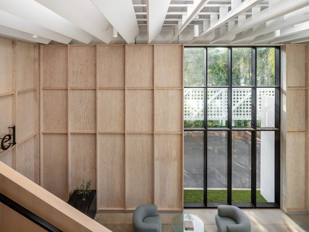 A bright contemporary interior viewed from a stair landing, featuring full-height light timber wall panelling, black-framed floor-to-ceiling glazing, and a partially visible seating area below. An exposed white structural ceiling with beams and services adds architectural interest, while a patterned breeze-block privacy screen and greenery are visible beyond the glazing.