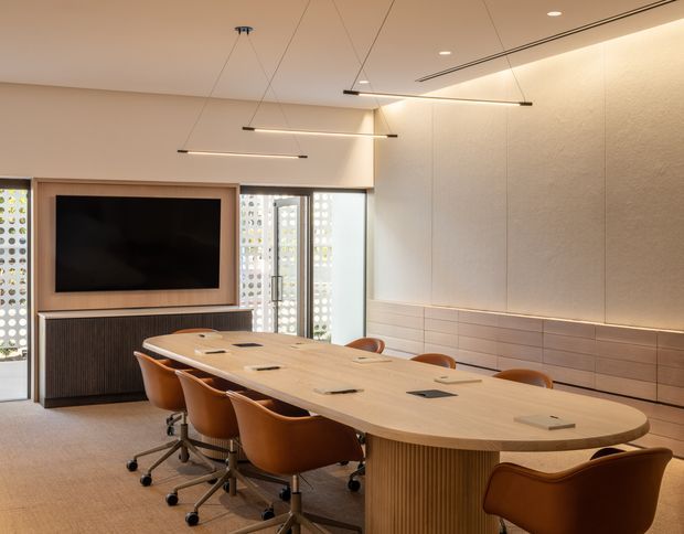 Contemporary meeting or dining room with a long rounded-edge timber table, integrated power outlets, curved tan leather office chairs, and a large wall-mounted screen set within built-in timber joinery. The room features warm neutral finishes, linear suspended pendant lighting, recessed ceiling lighting, textured wall panels, horizontal tiled wall detailing, and patterned privacy glazing.