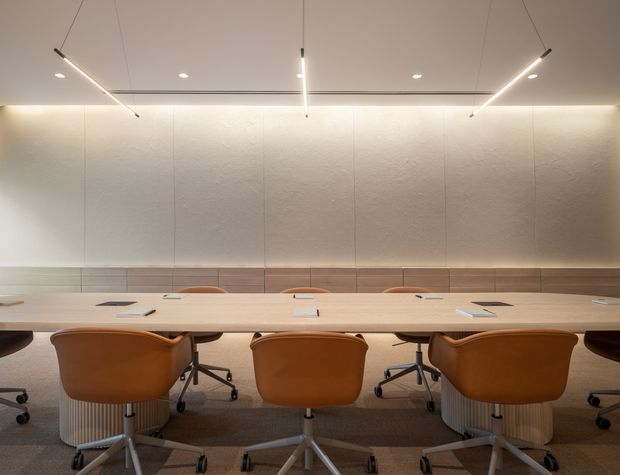 A refined contemporary meeting or boardroom with a long pale timber conference table, warm tan upholstered swivel chairs, textured light wall panels, a horizontal timber wall detail, and slim linear pendant lighting. The room has a calm, softly illuminated, minimalist corporate interior.