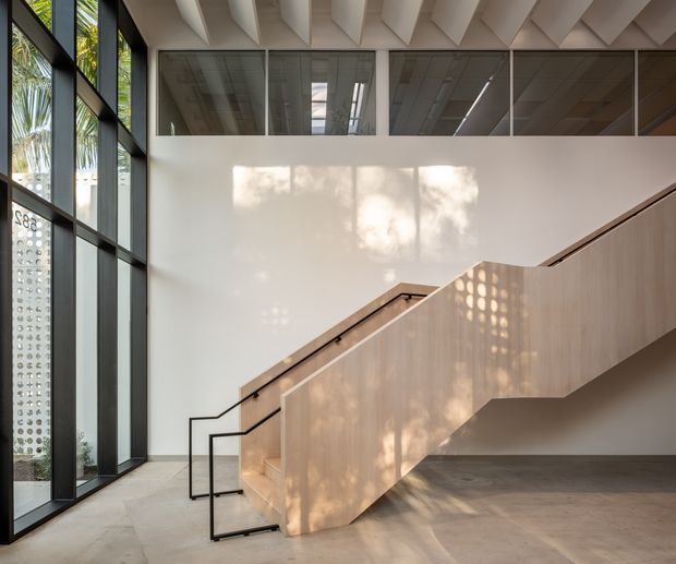 A refined contemporary stair hall with a sculptural timber-clad staircase, slim black metal balustrade, expansive black-framed glazing, and a high exposed-beam ceiling. Soft daylight and patterned shadows create a warm, airy architectural interior.