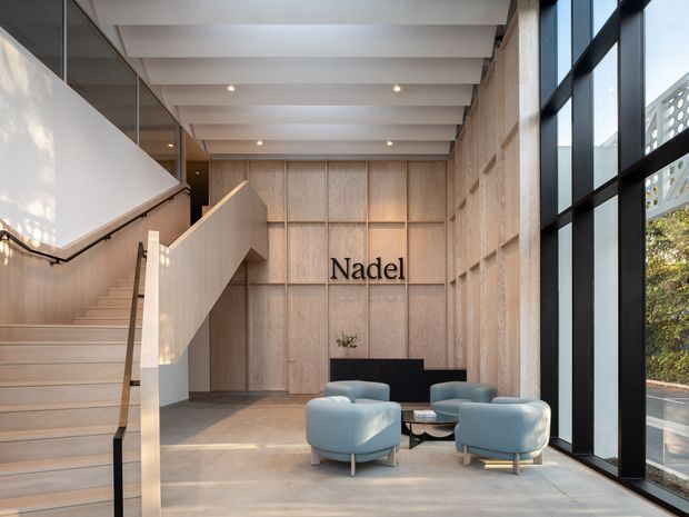 A refined commercial lobby or reception space with a prominent timber-lined feature wall bearing the “Nadel” sign, a sculptural staircase, exposed ceiling beams, polished concrete flooring, and full-height black-framed glazing. Pale blue lounge chairs and a low coffee table form a seating area beside the windows. The space has a warm, contemporary architectural character with abundant natural light.