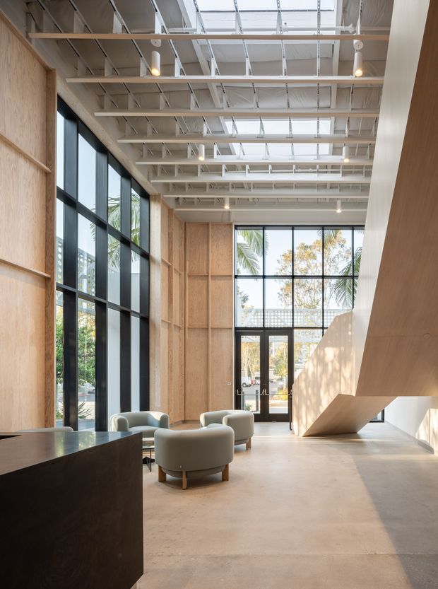 A bright, double-height contemporary interior resembling a reception or commercial lounge. The space features full-height black-framed glazing, pale timber or plywood wall panelling, exposed white structural roof framing, linear skylights, polished concrete flooring, sculptural pale upholstered lounge chairs, and a dramatic angular timber-clad stair or architectural feature. Natural light creates an airy, refined atmosphere.