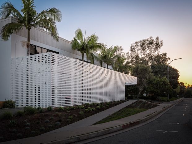 Exterior view of a contemporary low-rise building with a large white perforated masonry privacy screen wrapping the frontage. Palm trees are planted along the roofline and landscaped garden beds border a concrete pedestrian path beside the street. The scene is photographed at dusk, with clean horizontal building lines and a restrained white and grey palette.