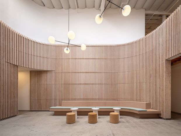 A spacious contemporary interior with a curved, floor-to-ceiling vertical timber-slat feature wall enclosing a built-in stepped bench. Three small cylindrical timber stools sit on a polished concrete floor. Sculptural globe pendant lights hang from a high exposed ceiling, creating a refined communal lounge or reception setting.