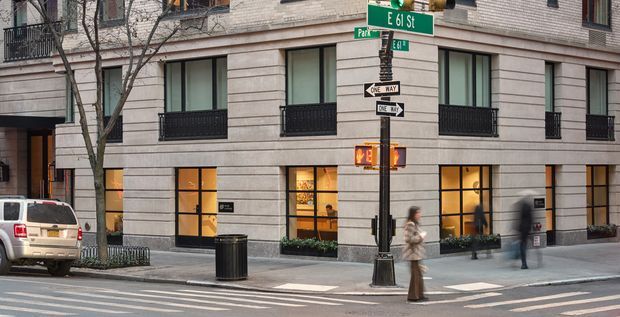 Street-level view of a multi-storey urban building on a corner, with a light stone or masonry facade, dark-framed windows, ground-floor illuminated commercial-style glazing, balconies, street signs, sidewalk, and crosswalk. The image primarily documents the building exterior and its urban streetscape.