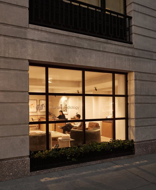 Exterior street-level view of a refined urban commercial facade with large black-framed glazed windows. Through the glazing, a warmly lit radiology reception and waiting area is visible, with two seated people, lounge chairs, a reception desk, and built-in cabinetry. The facade features horizontal stone or rendered bands, a dark metal balcony railing above, and a low planter at the window base.