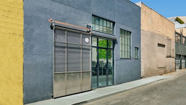 Street-level exterior view of a compact urban commercial or industrial-style building with a dark blue-grey rendered facade, large black-framed multi-pane windows, and a metal mesh security door. Adjacent weathered masonry walls and a narrow paved frontage reinforce the utilitarian urban setting.