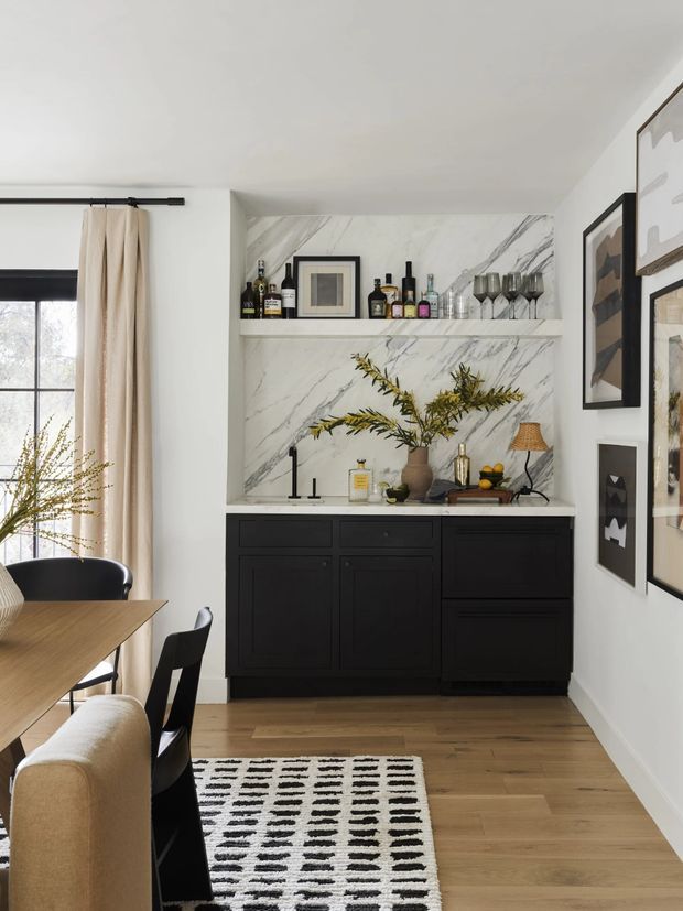 A refined dining and bar area with a black built-in sideboard, open marble-look shelving, bottles and glassware, framed artwork, and a timber dining table with black chairs. Pale walls, warm timber flooring, cream curtains, and a black-and-white patterned rug create a contemporary, high-contrast interior.