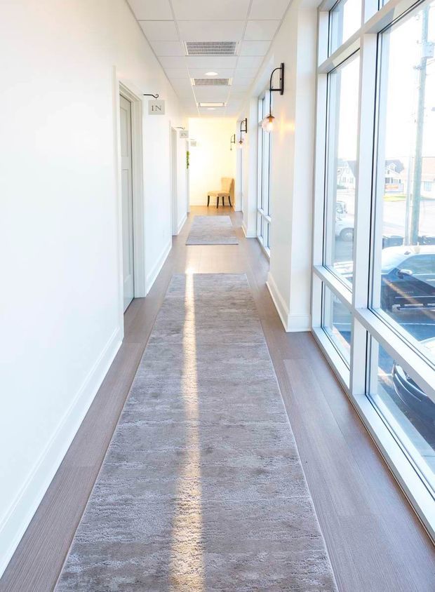 Bright, narrow commercial hallway with white walls, light wood-look flooring, a long grey runner rug, and full-height windows along one side. Multiple white paneled doors with small room-number plaques line the opposite wall, while black wall-mounted sconces provide warm accent lighting. A small seating area is visible at the far end, and parked vehicles can be seen outside through the glazing.