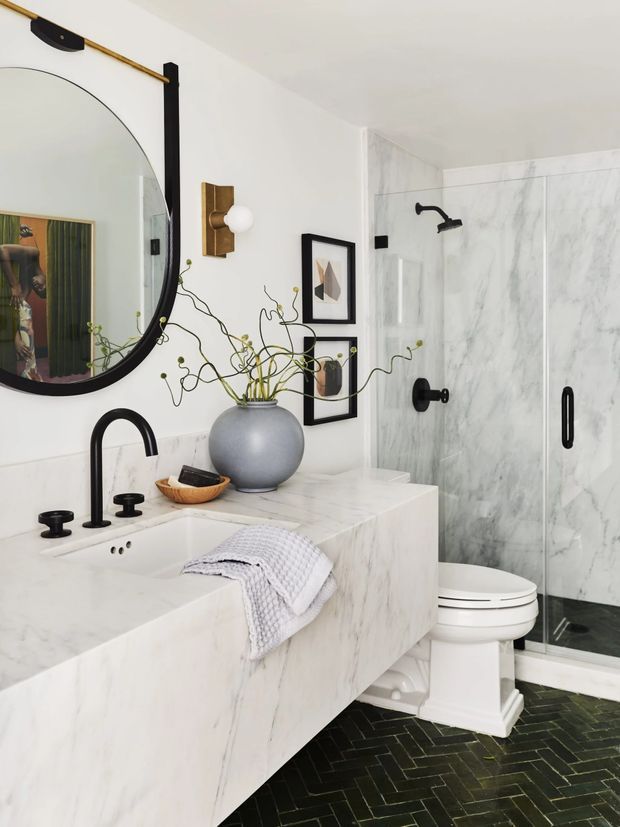A refined bathroom featuring a large white marble-look vanity with integrated basin, matte black tapware, a white toilet, and a glass-enclosed shower lined with veined marble-look wall panels. The floor has dark green-black herringbone tiles. A curved black-framed mirror, brass wall light, framed artwork, and a sculptural vase add decorative detail.
