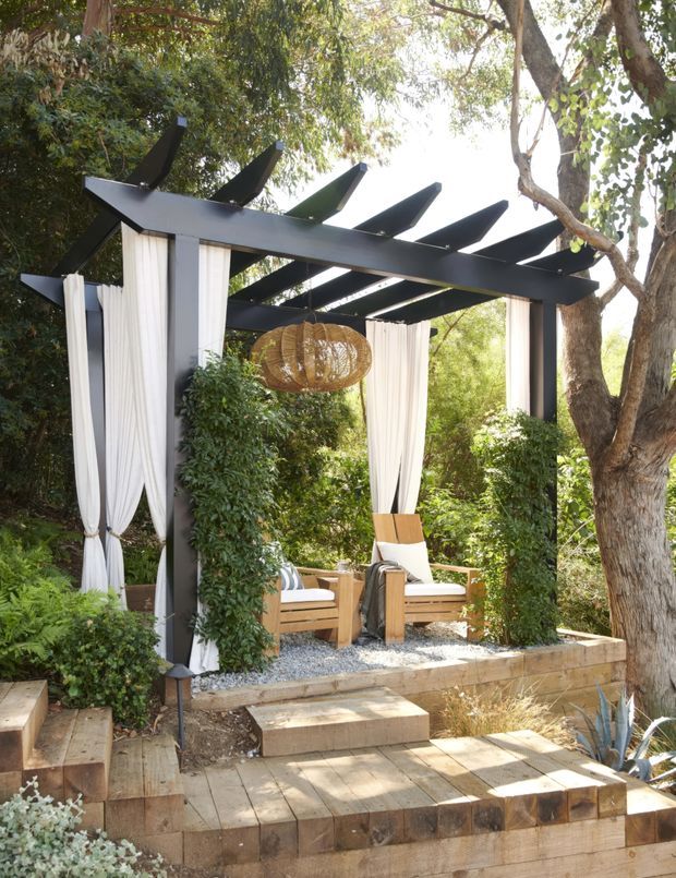 A landscaped outdoor lounge beneath a dark timber pergola with dramatic angled rafters. White curtains hang from the structure around timber-framed lounge chairs, with a woven pendant light overhead. The raised deck and steps are finished in warm timber, surrounded by dense leafy planting and mature trees.