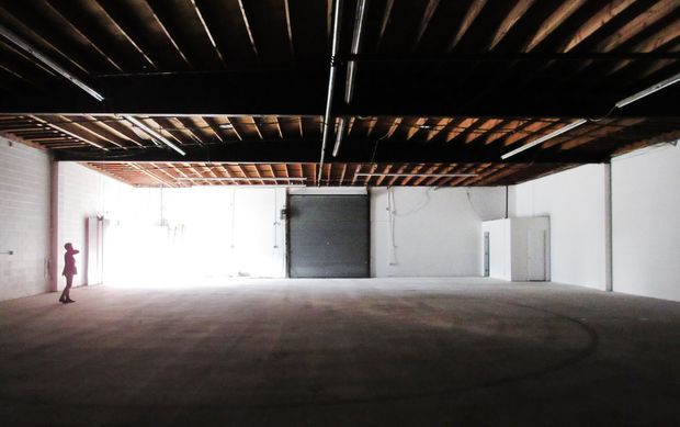 A large, empty industrial interior with a broad open concrete floor, exposed dark timber roof joists, visible linear services and fluorescent strip lighting, white-painted masonry walls, and a large roller door at the rear. A person at the far left provides scale. The space has a raw warehouse or studio character.
