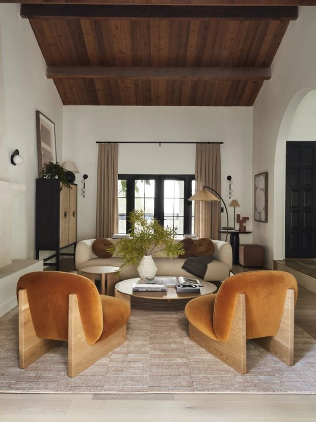 A warm, refined living room with a high exposed timber-beam ceiling, white plaster walls, dark-framed French doors, and an arched opening leading to a black paneled entry door. The room is furnished with a cream sofa, two sculptural rust-orange lounge chairs, a round coffee table, woven cabinetry, floor lamps, and a neutral textured rug. Natural light enters through the glazed doors, creating a cozy yet airy atmosphere.