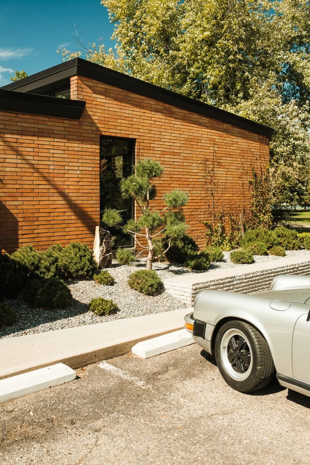 Exterior view of a low-slung brick building with a strong horizontal composition, flat roof, dark fascia, and a narrow glazed opening. The warm orange brick facade is bordered by gravel landscaping with clipped shrubs and a small sculptural tree. A paved parking area and partial vintage car appear in the foreground.