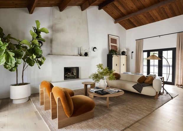 Warm, refined living room with a vaulted exposed timber-beam ceiling, white plaster walls, a minimalist fireplace, large black-framed glazed doors, and layered neutral furnishings. The room includes a cream sofa, two sculptural ochre velvet lounge chairs, a round timber coffee table, textured area rug, tall indoor plant, and built-in or freestanding storage. Natural light creates an inviting, contemporary atmosphere.