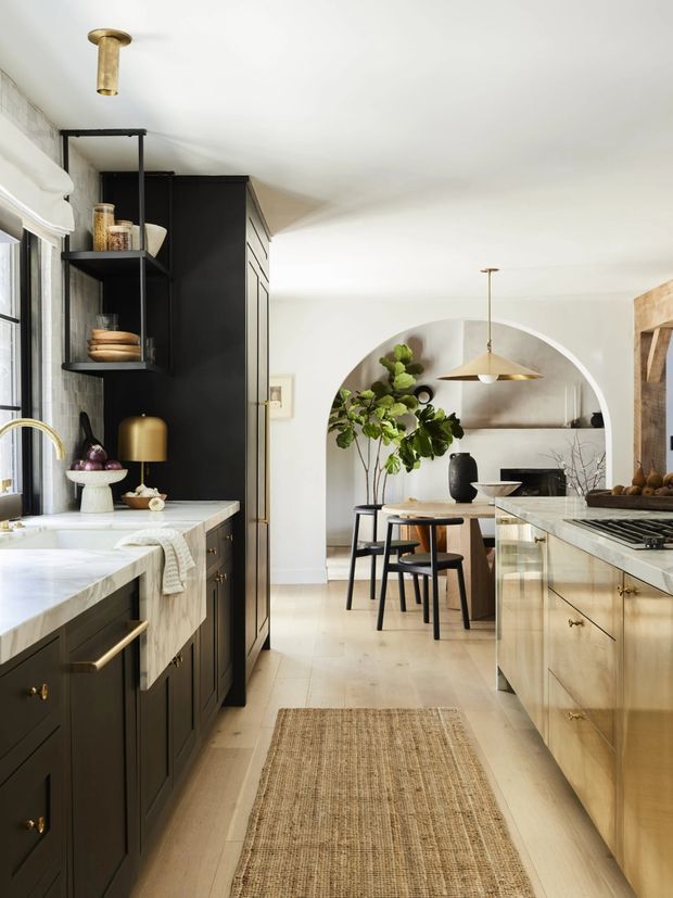 A refined galley kitchen with dark charcoal cabinetry, pale stone benchtops, brass hardware and a timber-toned island. The kitchen opens through a large arched passage to a dining area with a round table and stools, while open shelving, pendant lighting, a large window and warm natural styling create an inviting, layered interior.