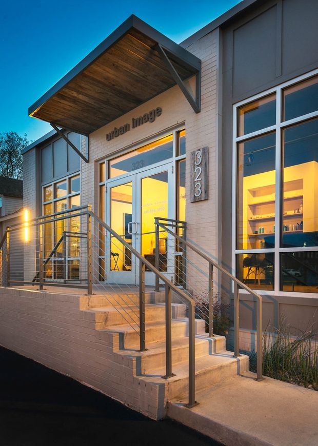 Dusk exterior view of a contemporary commercial building entrance. The facade combines light painted brick with dark vertical panel cladding and large aluminium-framed windows. A projecting timber soffit canopy shelters glazed double entry doors, with illuminated interiors visible through the windows. Concrete steps, an accessible-looking ramp or raised entry approach, and simple metal cable railings lead to the entrance, which is marked with the number 323 and the words “urban image.”