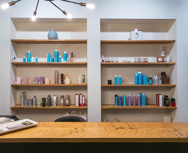 A retail-style interior with two large recessed wall shelving units displaying neatly arranged bottles, boxes and beauty products. Warm timber shelves and a substantial timber counter contrast with light painted walls, while a black multi-arm pendant light hangs overhead. The space has a clean, curated, contemporary feel and appears to be a beauty salon or product display area.