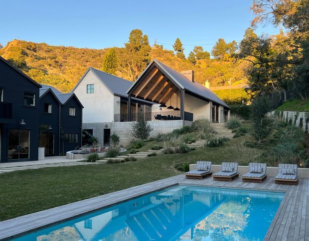 A contemporary hillside residential property with a vivid rectangular swimming pool in the foreground, surrounded by a timber deck and three loungers. The main house features steep pitched gable roofs, dark vertical cladding on one wing, pale rendered walls, extensive glazing, and a dramatic open gable entertaining area. Native-style grasses, tiered landscaping, and wooded hills form the setting.