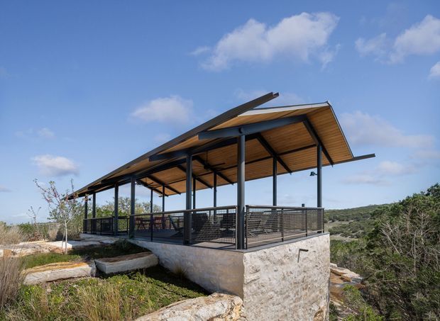 Elevated open-sided pavilion or outdoor living structure set on a rocky, vegetated site. It features a broad asymmetrical pitched roof with warm timber soffit lining, slender dark steel posts and beams, dark mesh balustrades, and a rough stone or concrete masonry base. The structure overlooks a natural landscape beneath a clear blue sky.
