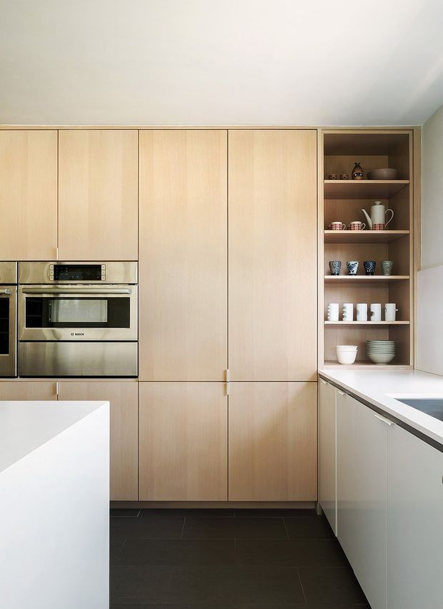A clean, contemporary kitchen featuring a full-height wall of pale timber cabinetry, integrated stainless-steel ovens, and an open display shelving niche with crockery. A white island and peninsula-style benchtop frame the foreground, while dark large-format floor tiles provide contrast. The space has a simple, refined fit-out with concealed storage and a neutral palette.