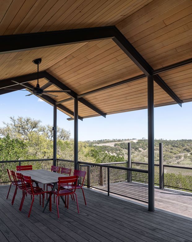 A spacious covered outdoor deck or terrace with dark timber decking, a warm timber-lined ceiling, and black steel posts and beams. A ceiling fan hangs above an outdoor dining table with red chairs, while mesh balustrades frame views across a dry, wooded landscape. The open-sided structure feels suited to entertaining and outdoor dining.