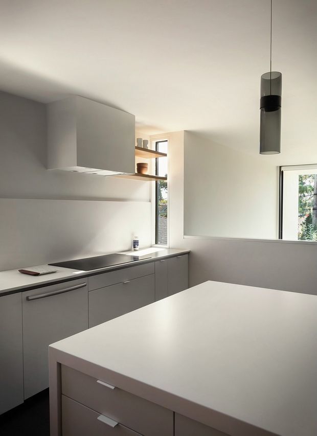A bright, minimalist contemporary kitchen with grey handleless cabinetry, a dark induction cooktop, pale solid-surface island, white walls, open timber shelving, and slim black-framed windows. A cylindrical dark pendant light hangs above the island, with greenery visible outside.