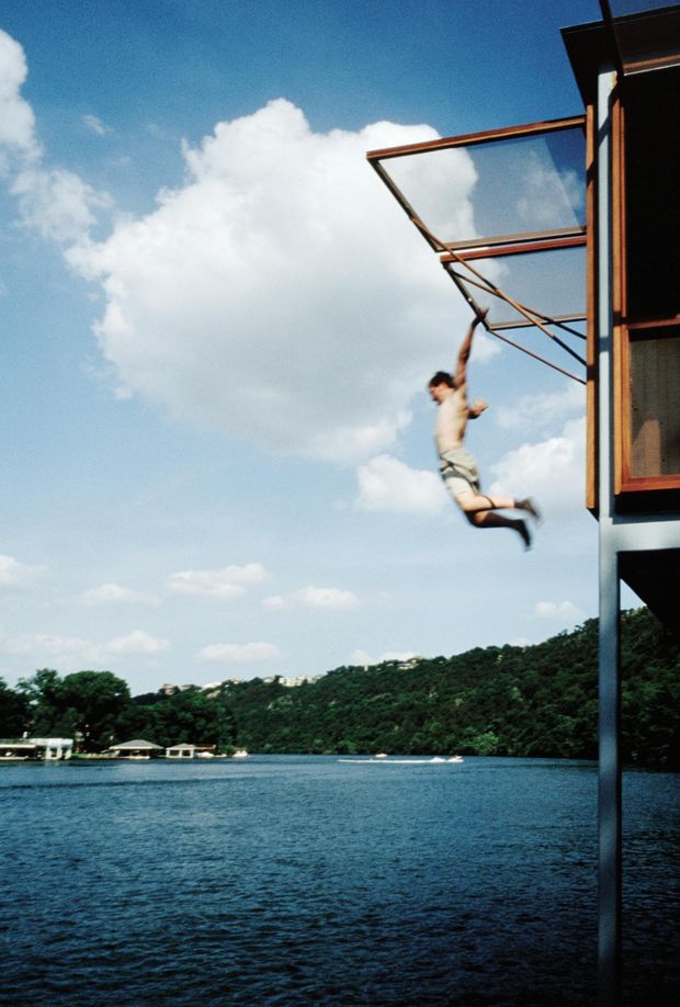 A waterside modern structure viewed from outside, with a person swinging or jumping from a projecting glass-fronted platform. The building has dark metal structural supports, timber-toned framing, and large glazed openings, set above a broad body of water with a densely wooded opposite shoreline under a bright blue sky.