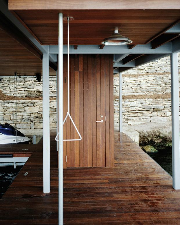 Covered timber deck and dock-like exterior walkway beside a rustic stacked-stone wall, with a vertical timber-clad door, exposed grey steel framing, timber soffit, and a suspended industrial-style light. A small boat is partially visible at the left edge, suggesting a waterside setting.