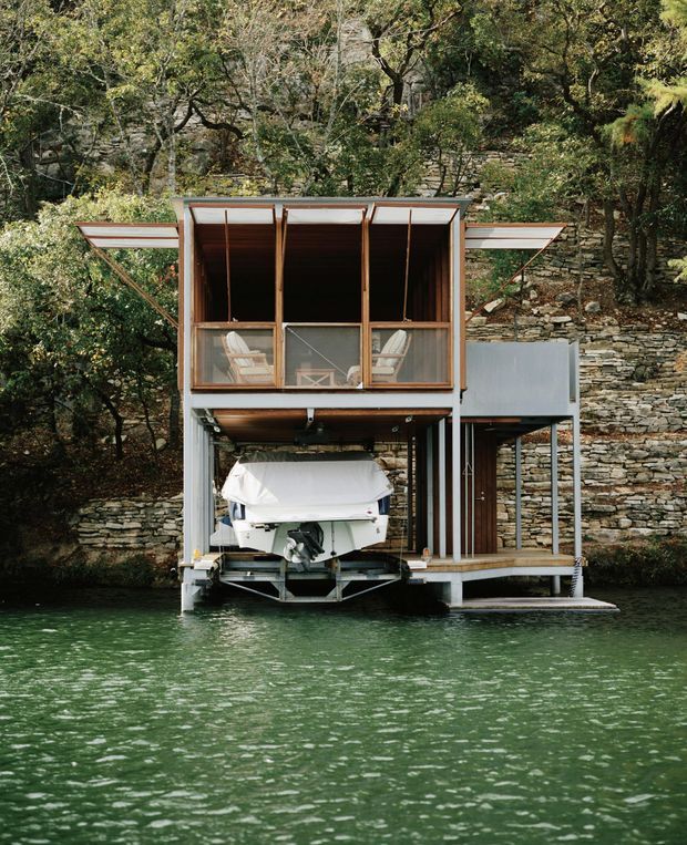 A compact waterside boathouse set directly above a green body of water, with a covered boat storage bay at ground level and an enclosed upper-level seating or living space. The structure has extensive timber-framed glazing, projecting wing-like roof or awning elements, grey structural panels, and a small side landing. Behind it is a steep, heavily wooded site with layered stone retaining walls. The image is an exterior architectural photograph viewed from across the water.
