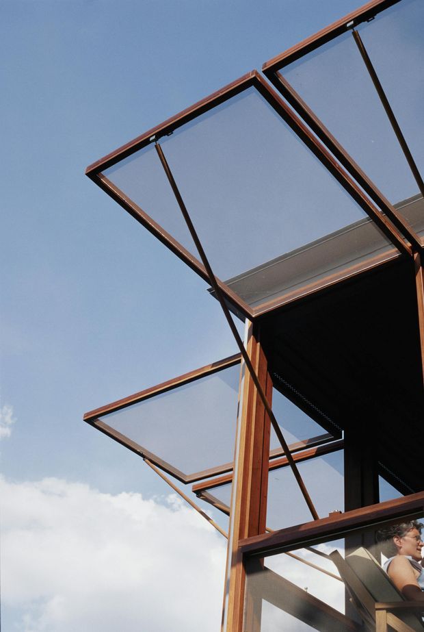 Upward-looking exterior architectural detail showing a series of large glass awning or roof panels held in dark timber frames with slender metal support rods. The glazing projects from a modern structure against a blue sky, with part of the building facade and a person visible at the lower right.